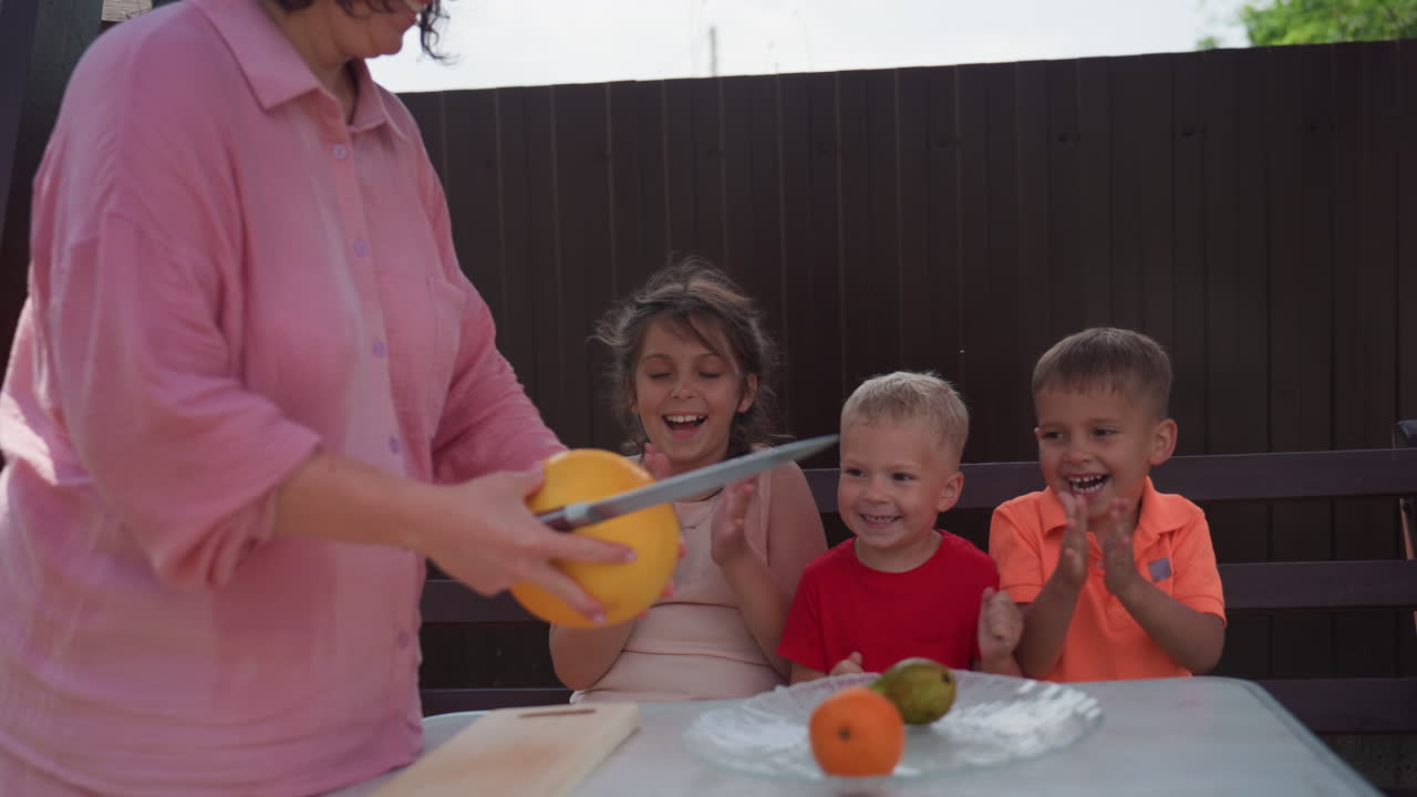Bambini che guardano un adulto tagliare frutta fresca a un tavolo in giardino, volti emozionati e condivisione dell'attesa, atmosfera da picnic con altalena e giardino verde, momento di snack sano e gioia di gruppo