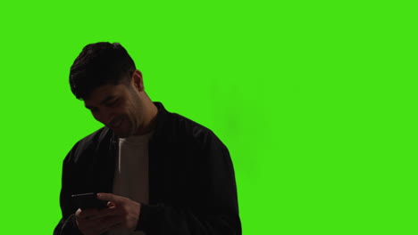 Young-Man-Writing-And-Sending-Text-Message-On-Mobile-Phone-Standing-Against-Green-Screen-Background-With-Low-Key-Lighting