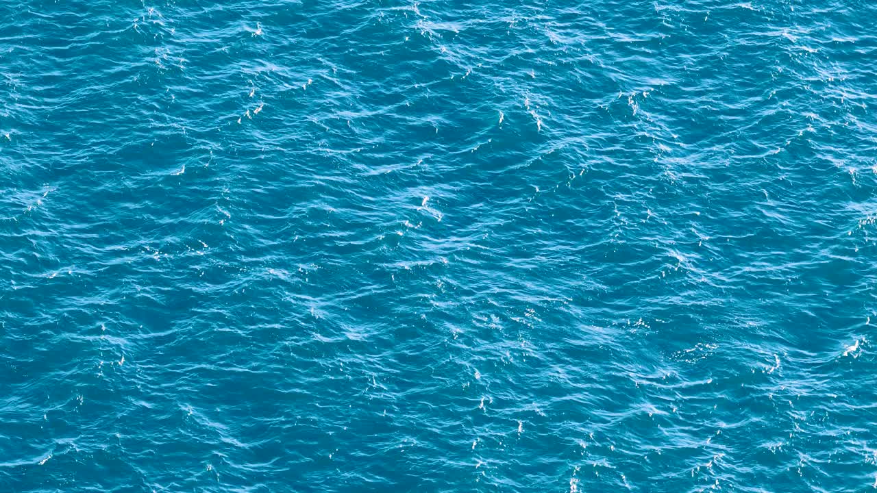 Gentle ocean waves ripple across a vibrant blue sea, captured from above in bright daylight with subtle camera movement and natural lighting
