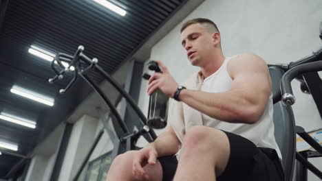 young athlete seated on weight bench grabbing water bottle inspecting gym during post workout rest between intense chest press sets catching deep breath with towel draped over shoulder muscular arms