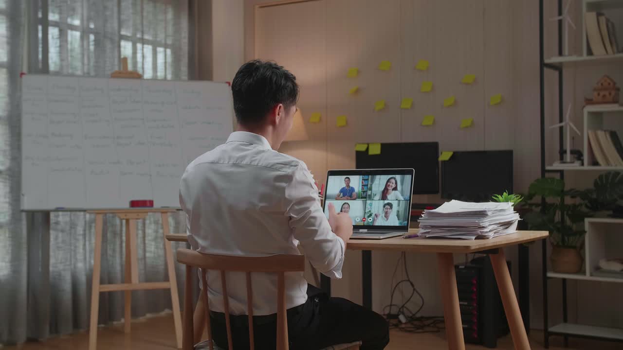 オフィスで書類作業をしながらノートパソコンでビデオ通話をするアジア人男性の後ろ姿。