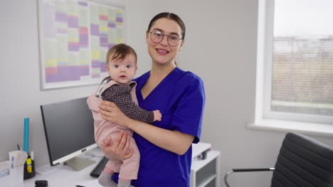 Portrait-of-a-confident-and-happy-brunette-doctor-girl-in-glasses-and-a-blue-uniform-who-is-holding-a-little-baby-girl-in-her-arms.-Portrait-of-a-confident-and-qualified-female-doctor-in-a-blue-pediatrician-uniform-in-a-modern-clinic