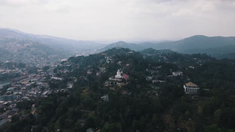 big white bahirawakanda vihara buddha statue on one of the hilltops among the beautiful mountains of green nature in sri lanka with kandy city in the valley on a cloudy day. aerial drone panning shot