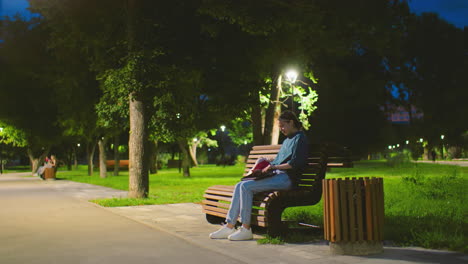 young woman sits on bench in evening, in tranquil park, opening her laptop, background includes lush greenery, trees, a glowing light pole, and people casually strolling