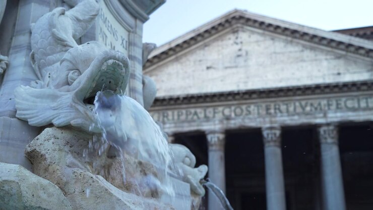 Primo piano della scultura di pesce della Fontana del Pantheon che getta acqua, con le colonne del Pantheon leggermente sfocate sullo sfondo.