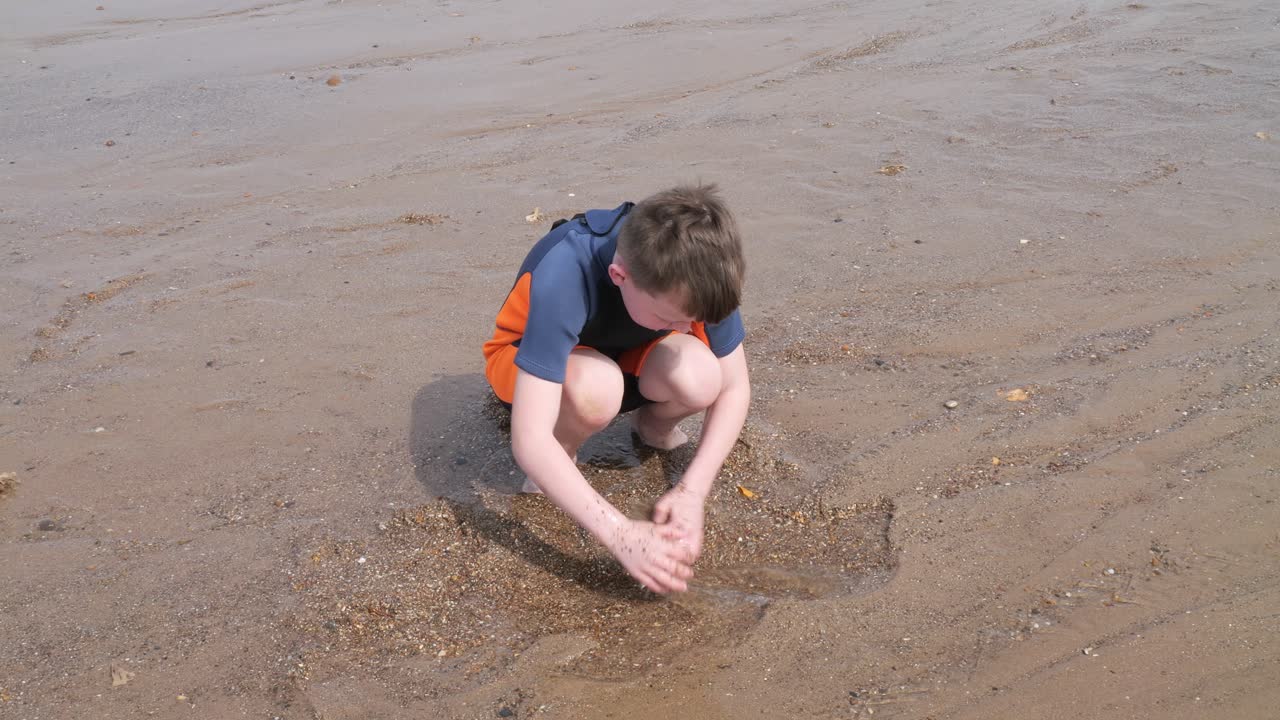 Young boy in a wetsuit on a beach digging in the sand. Playing with a surfboard and practicing skimming on the water. Seaside scene. Ocean waves breaking on the beach. Holiday time season