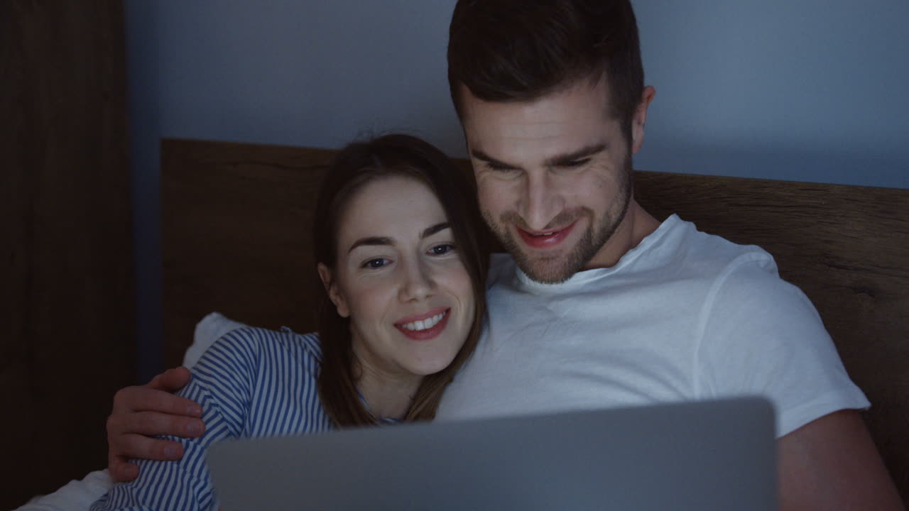 sonriente pareja joven caucásica de buen aspecto viendo algo divertido en la computadora portátil en la cama por la noche