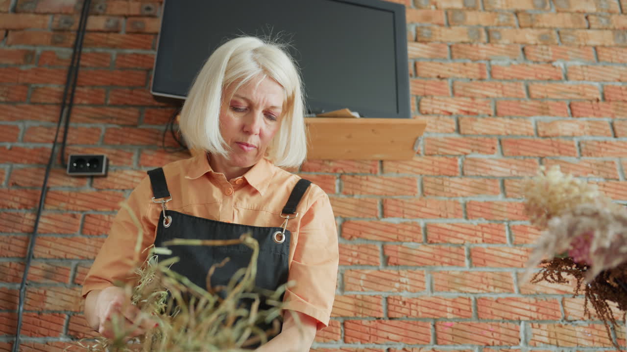 Decorator with blonde hair in black apron arranging natural hay and dried flowers on wooden table against brick wall, working on handmade rustic wreath creation with tools and materials in cozy workshop