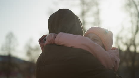 a little girl in a pink cap and jacket being carried by a man wearing a brown jacket, as he turns her around and she holds him tightly