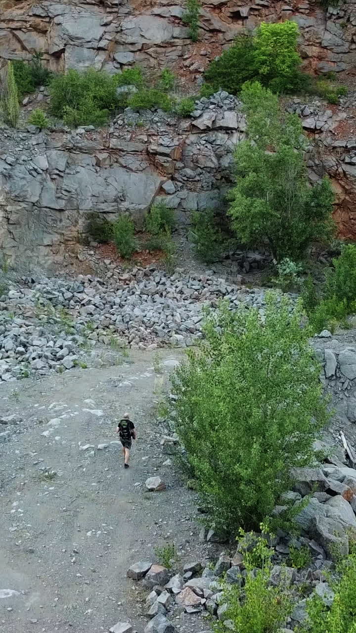 Traveler with a backpack is walking near the river on the stone background in summer. A tourist male hiking through the canyon with a wonderful landscape. Aerial view Vertical video