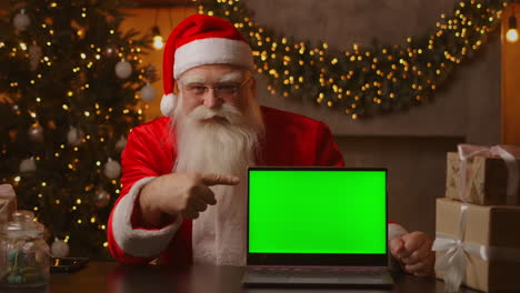 In-the-background-of-the-Christmas-tree-an-elderly-Santa-Claus-is-holding-a-laptop-and-pointing-at-the-screen-with-a-chromakey.-Laptop-with-a-green-screen.-Santa-looks-at-the-camera.-High-quality-4k-footage