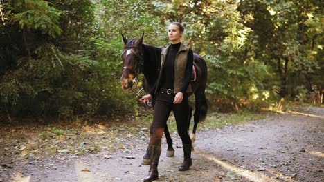 slow motion shot: beautiful young female jockey is walking with a stunning brown horse with white spot on forehead in the forest