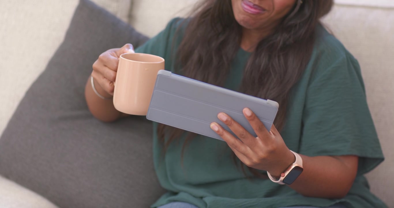 Glancing at tablet, adult African American woman sipping mug, adjusting tablet on couch and reading