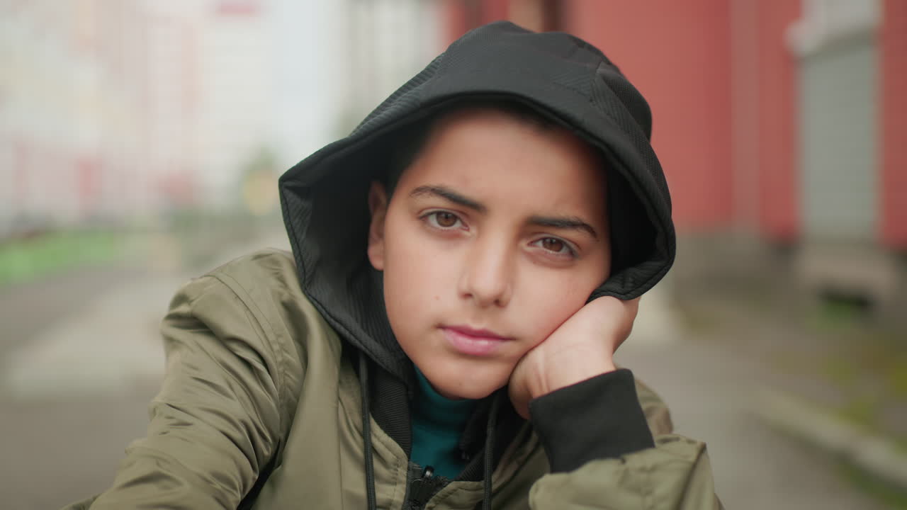 Young boy in hooded jacket sitting outdoors, resting head on hand thoughtfully, gazing forward with calm and reflective expression, blurred urban background