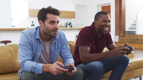 two male friends sitting on sofa in lounge playing video game