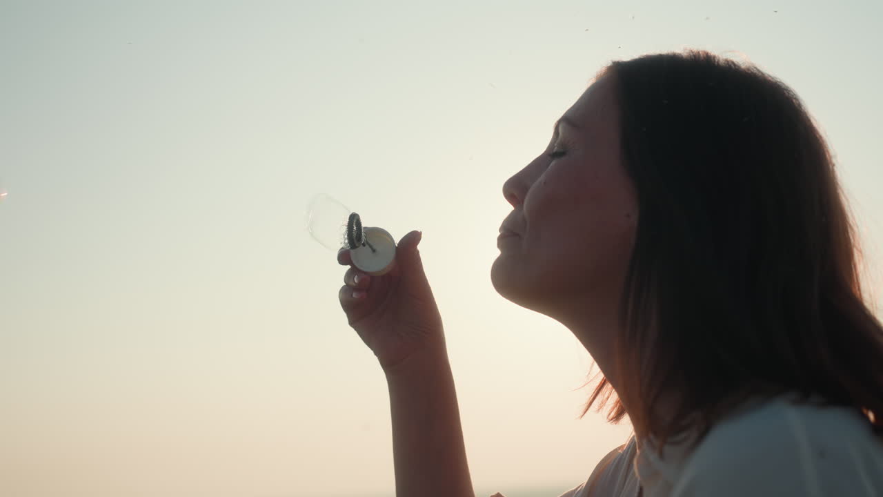 Close up side view of woman gently blowing bubbles with closed eyes under warm sunset glow, calm expression on face, serene atmosphere with soft light and floating particles
