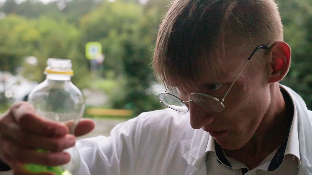 Close view of botany student in white coat sitting outdoors chewing snack and sipping lemonade drink while looking aside with relaxed expression during break and wet pavement in blurred background