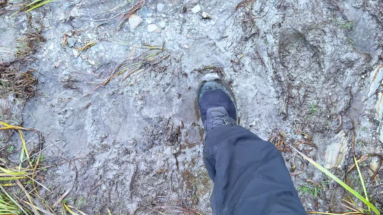 Person walks along muddy, uneven trail with wet grass, overcast daylight, and handheld camera movement