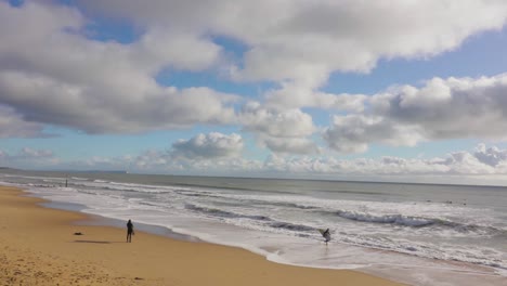 A-lone-surfer-stands-watching-the-waves,-beach-and-partly-cloudy-sky-in-the-background