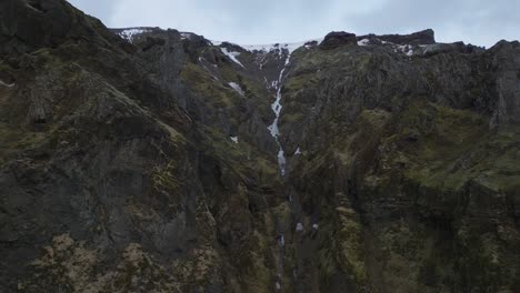 Aerial-landscape-view-of-a-waterfall-flowing-down-icelandic-mountain-cliffs,-melting-snow,-on-a-moody-day