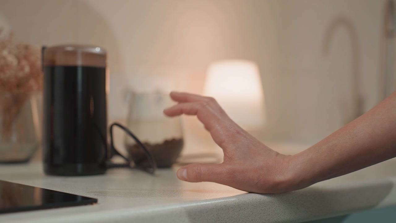 Hvite kvinnefingre trommer på kjøkkenbenken under varmt lampelys, kaffepresse og glasskrukke synlig i bakgrunnen, fingertuppene spiller en myk rytme som en melodi, nærbilde av håndbevegelser formidler romantikk