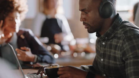 Joven-Afroamericano-Usando-Una-Computadora-Portátil-En-Un-Café-Navegando-En-Línea-Revisando-Mensajes-De-Correo-Electrónico-Disfrutando-Bebiendo-Café-Escuchando-Música-Trabajando-En-Un-Restaurante-Ocupado