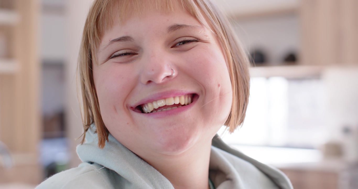 Female reacting to cue shifting soft smile into broad laugh tilting head in kitchen wearing hoodie