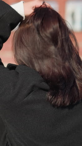 vista trasera de cerca de una joven de cabello marrón en un abrigo negro ajustando su cabeza, mostrando tres anillos en su mano izquierda, en una calle urbana soleada