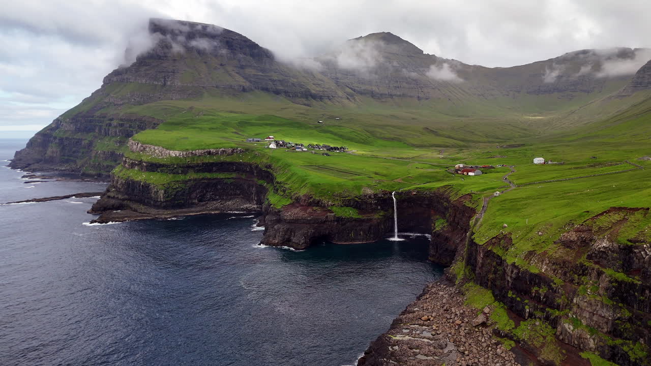 Aerial view of Gásadalur waterfall plunging into the Atlantic Ocean, framed by dramatic cliffs, lush green Faroe Islands landscape, and traditional village houses
