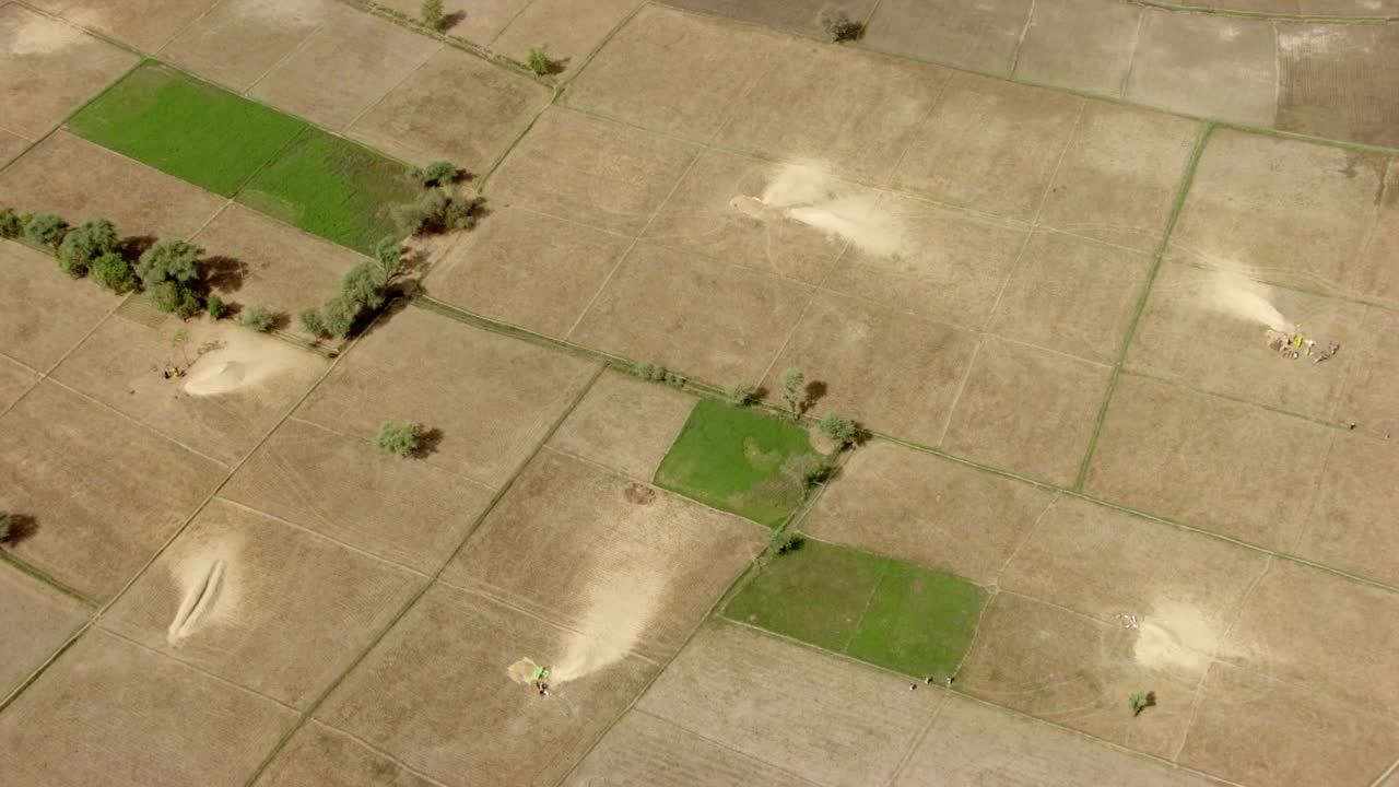 Drone footage captures a wide aerial view of agricultural fields divided into rectangular plots. Most plots appear dry and barren, while a few show green vegetation, indicating active cultivation