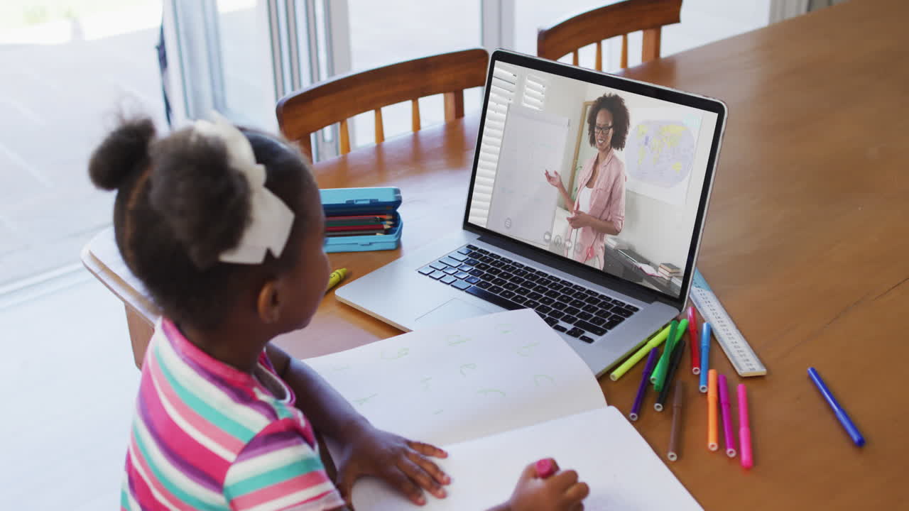 African american girl doing homework while having a video call with female teacher on laptop at home