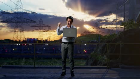full body of an asian male professional worker standing with his laptop near high voltage tower, industrial facility, he raises his fist up with screaming goal