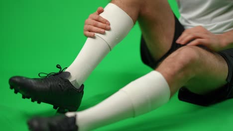 unrecognizable caucasian football player with ache in leg sitting on green screen. young footballer with painful limb at chromakey background. sport accident and trauma concept.