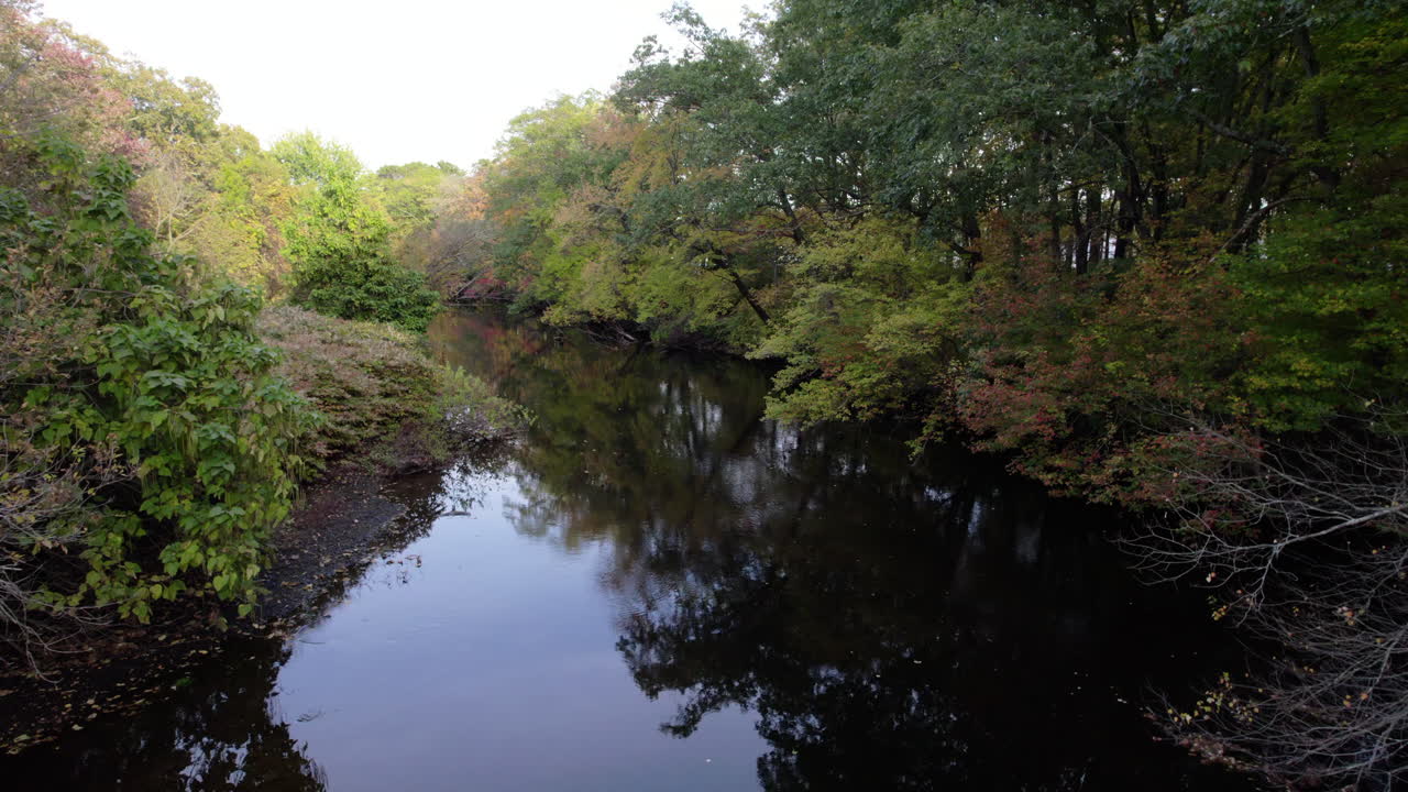 Aerial drone video of the peaceful Pawtuxet River in Cranston, Rhode Island lined with color changing fall foliage and bookended by a railroad bridge and a road bridge