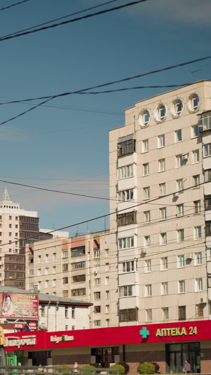 Calle de la ciudad con edificios de apartamentos, escaparates, transporte. Escena diurna bajo un cielo azul despejado con cables entrecruzados y líneas de cable, bloques residenciales de varios pisos, toldos comerciales rojos, autobús que pasa