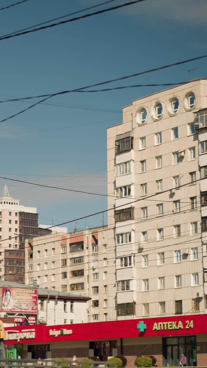 Strada cittadina con palazzi, vetrine, mezzi pubblici. Scena diurna sotto un cielo azzurro limpido con cavi e linee elettriche che si incrociano, edifici residenziali a più piani, tende commerciali rosse, autobus di passaggio
