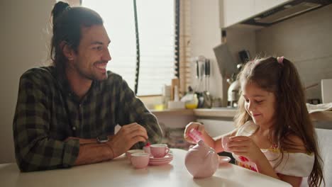 Happy-brunette-man-in-green-checkered-shirt.-Lonely-father-plays-tea-party-game-with-his-little-daughter-brunette-girl-in-pink-dress-using-toy-pink-cups-and-kettle-in-kitchen