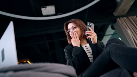 young woman wearing glasses looks at laptop with surprise. low angle view on the brunette with smartphone finding out shocking news.