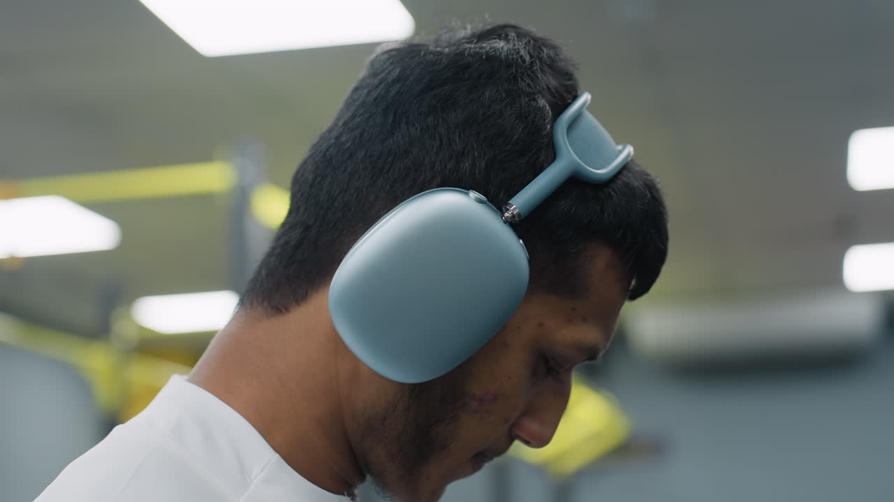 close up side view of young man adjusting over ear headphone strap while glancing down in bright gym environment, blurred passerby and yellow workout machine in background