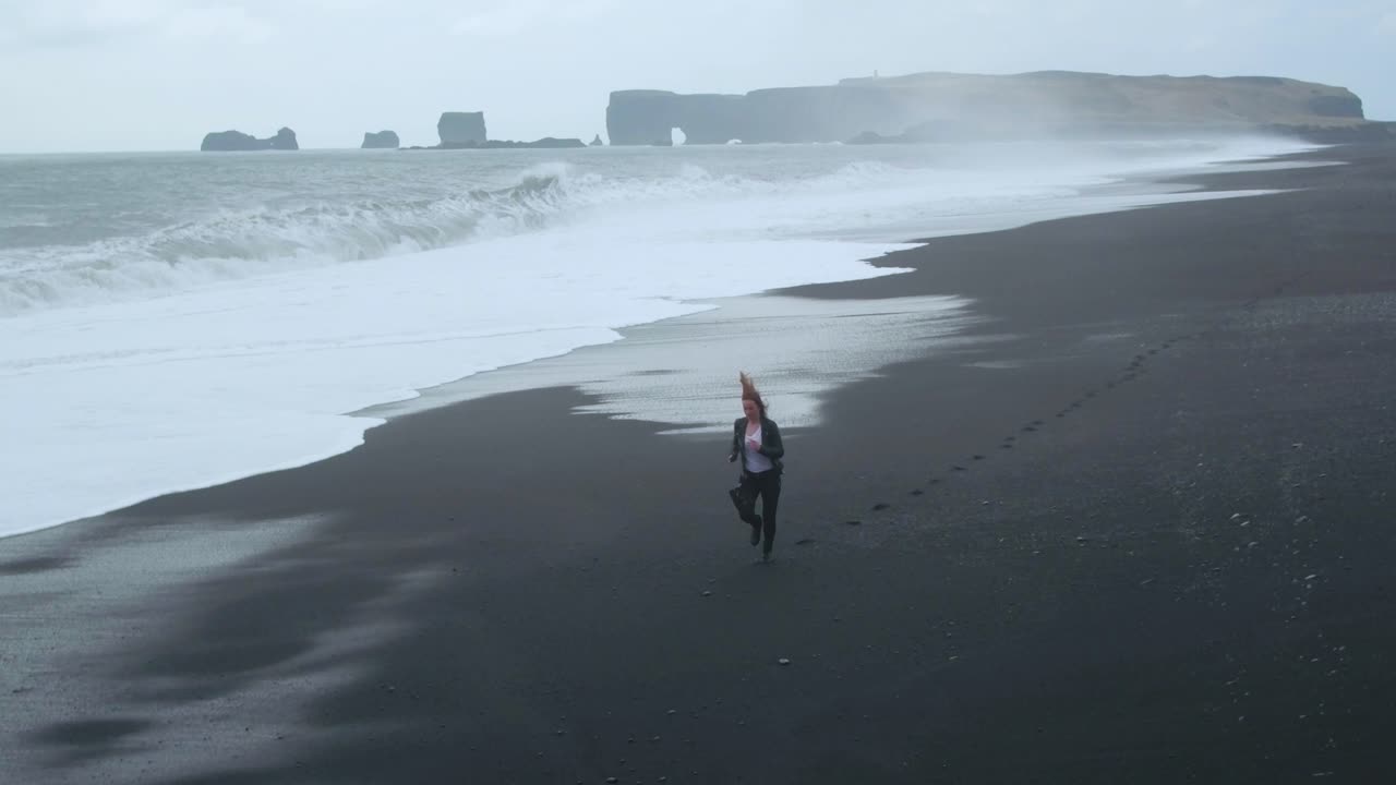 Woman running on black sand beach in rough sea day. Aerial flying backwards