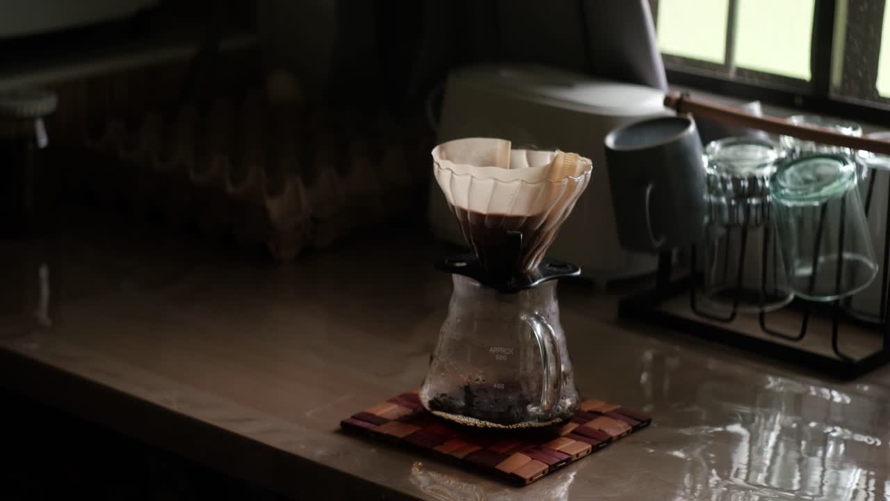 Medium static shot of Gooseneck kettle pouring hot water into V60 dripper and filter paper for coffee pour-over brew over glass carafe during morning