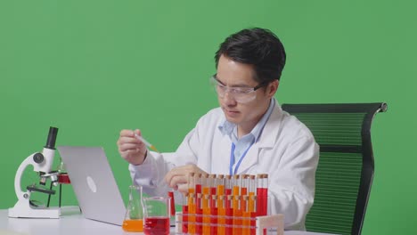 side view of asian man scientist making experiment with test tube and typing on a laptop while working on the table with microscope in the green screen background laboratory