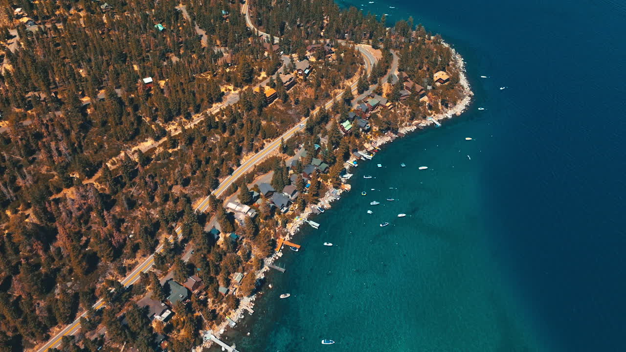 Flying over the rocky shore of Lake Tahoe overgrown with pine trees and built up with cottages. Beautiful water azure with some boats sailing on. Top view.