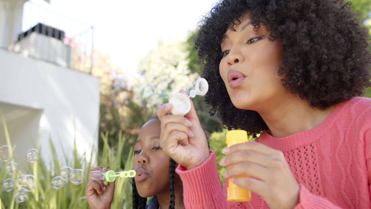 Happy african american mother with daughter blowing bubbles in garden at home, slow motion