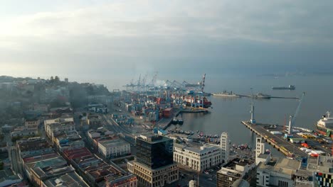 aerial jib up over buildings near cranes, containers and ships docked in valparaiso sea port on a cloudy day, chile