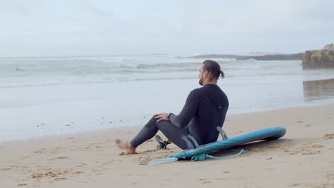 back view of a tired sportsman with artificial leg sitting on sandy beach after surfing workout in ocean, looking at waves and relaxing