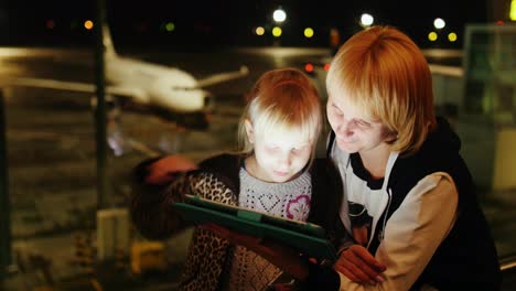 mamá e hija juegan en la tableta en el aeropuerto a última hora de la tarde, el avión es visible fuera del