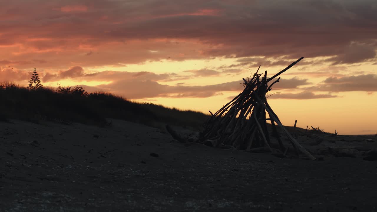 Fiery orange sunset, golden sky over teepee of wooden sticks on beach