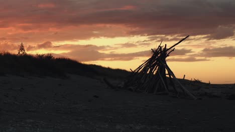 fiery orange sunset, golden sky over teepee of wooden sticks on beach