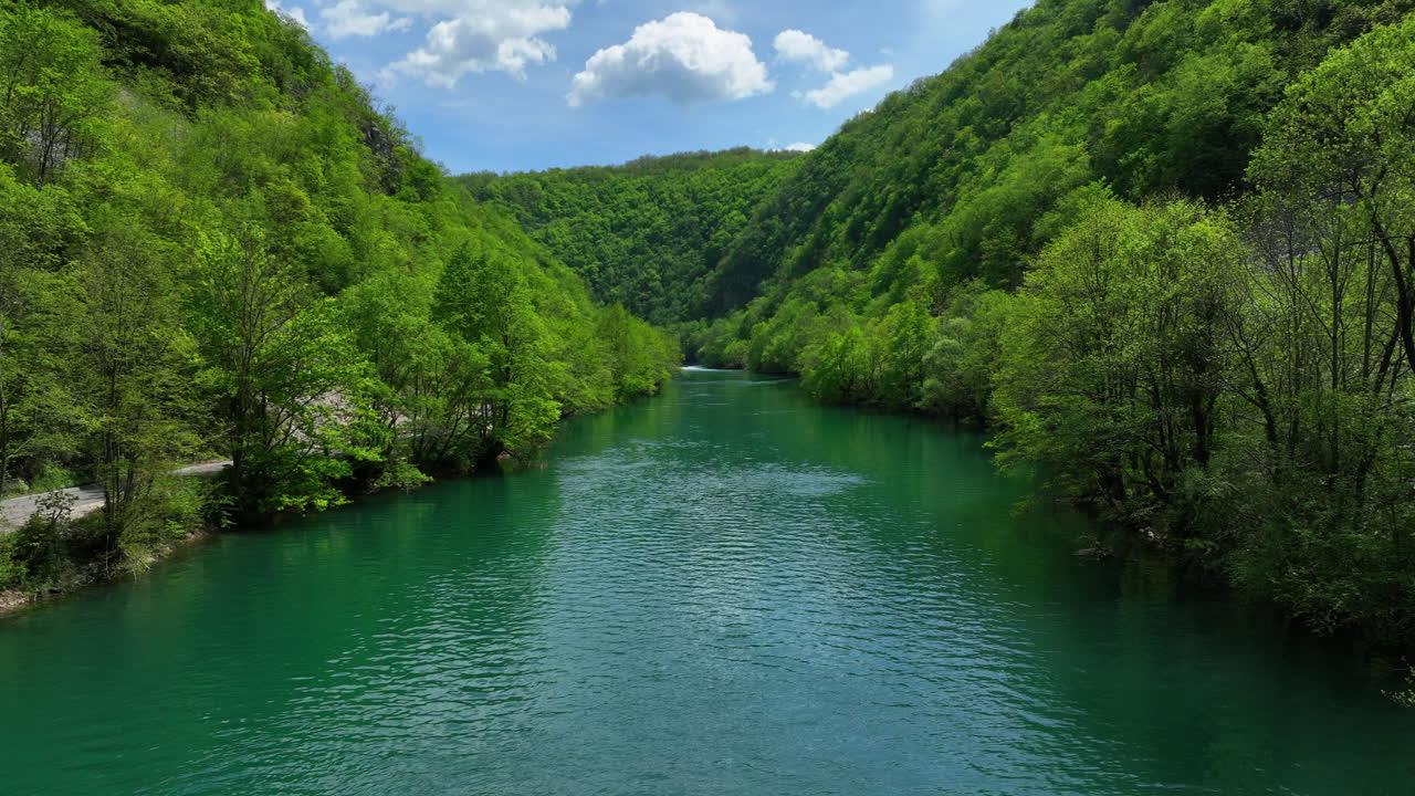 Ripresa aerea bassa di un fiume verde e limpido circondato da alberi rigogliosi e appena germogliati all'inizio della primavera, sotto un cielo con nuvole sparse